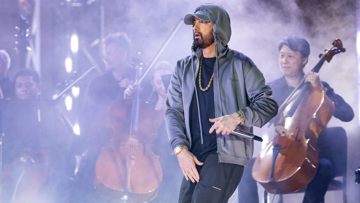 Eminem performing on stage wearing a hooded jacket and gold chain, with a live orchestra in the background