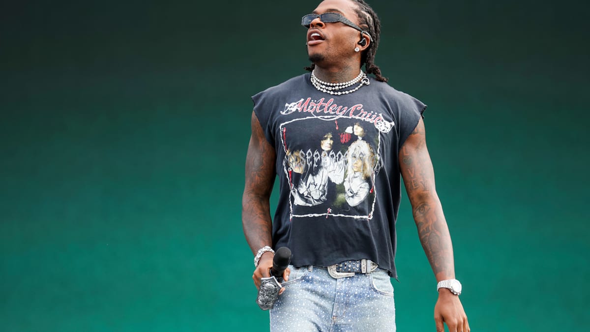 Rapper Gunna performs on stage, wearing a sleeveless Motley Crue t-shirt, sunglasses, and sparkly jeans, holding a microphone