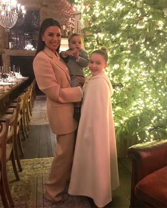 a women wearing a pink suit holds a baby as a girl stands next to her wearing a cape with a long dining table in sight near a window