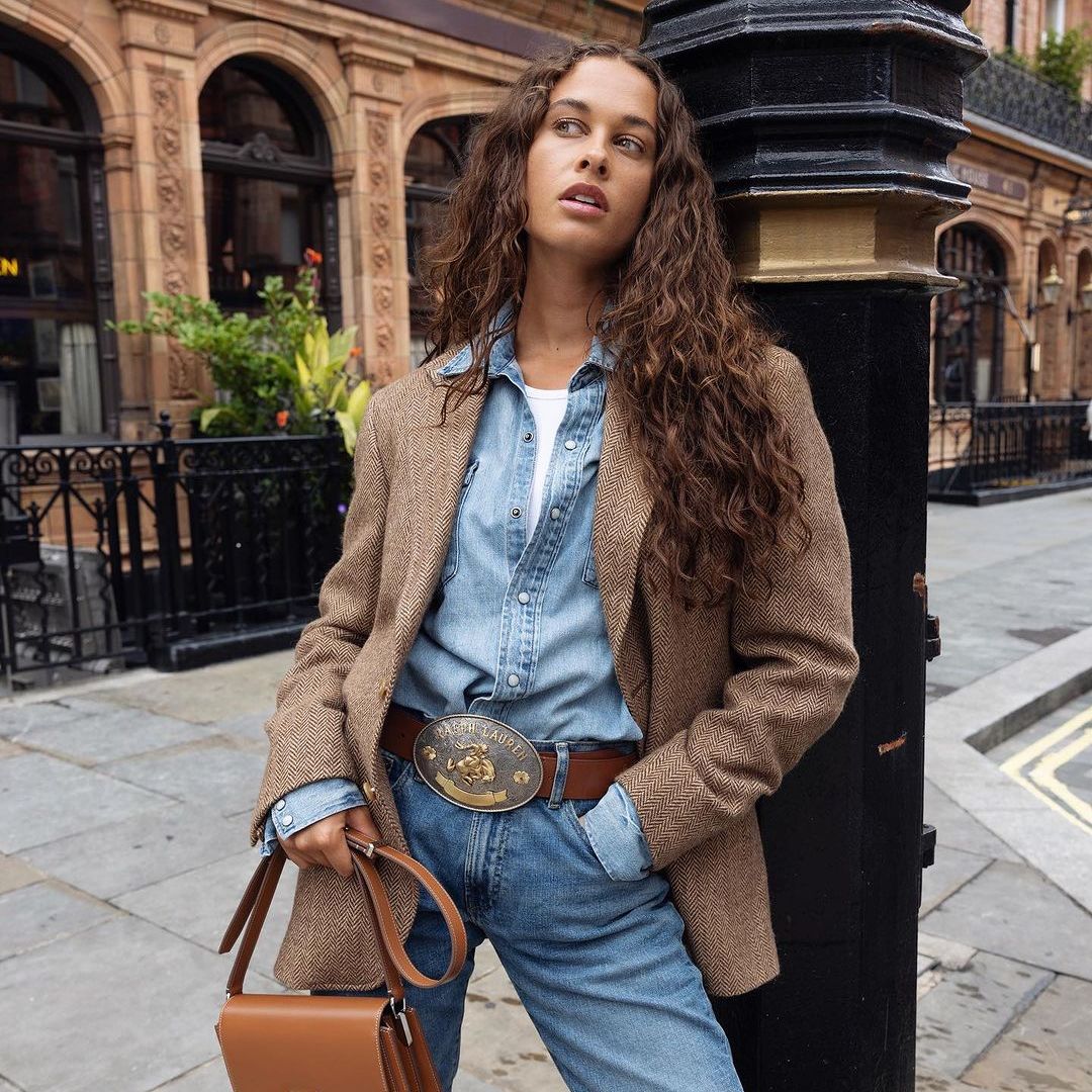 Sarah Lysander wears jeans, a denim shirt, blazer and belt buckle to pose against a street light in london