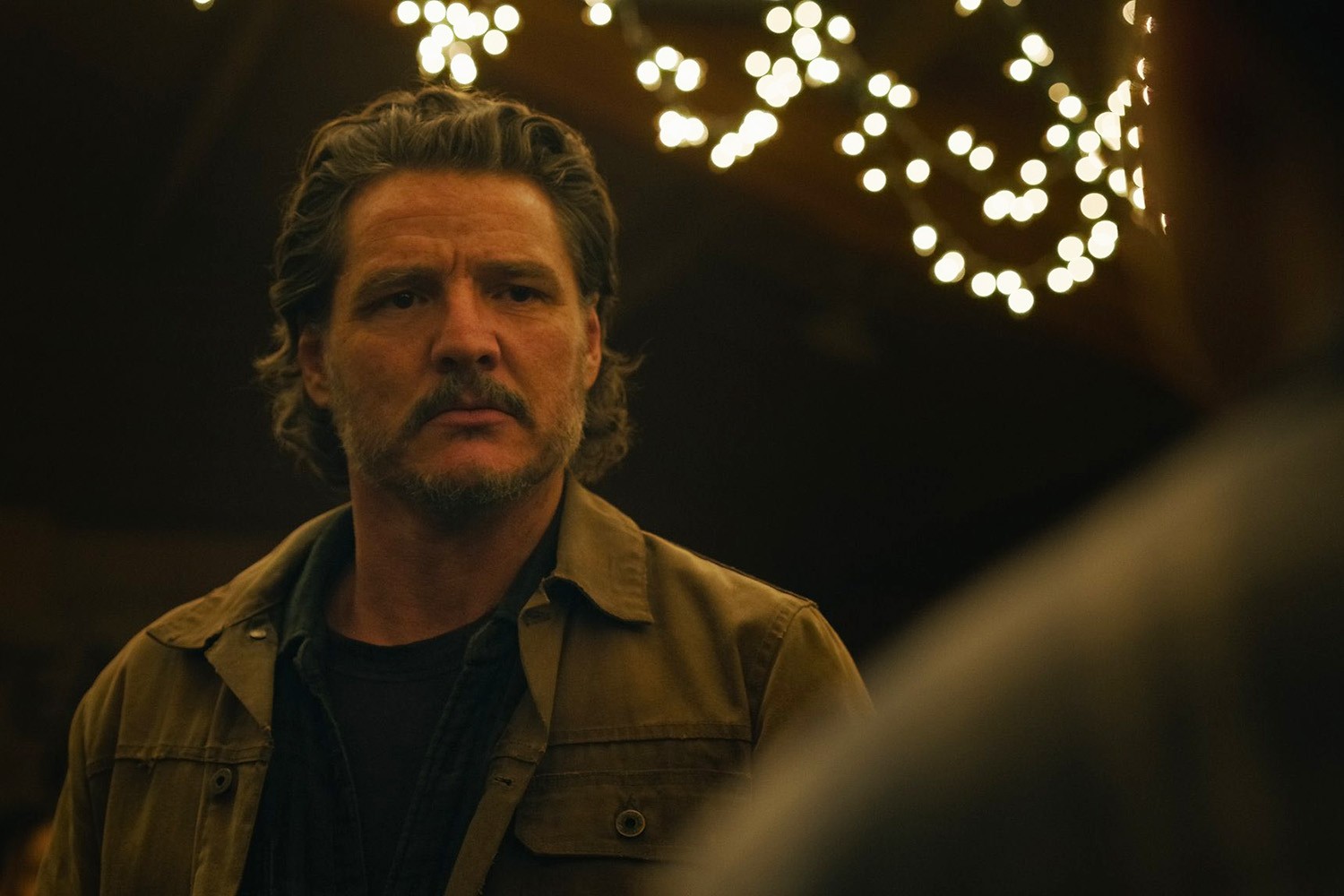 Joel (Pedro Pascal) in The Last of Us season 2, standing in a dimly lit area with fairy lights in the top right-hand corner