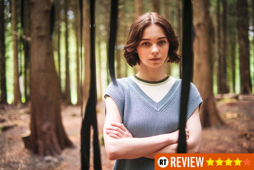 Pip standing in the woods with her arms crossed, looking directly into the camera. There's an orange RT 4-star banner in the bottom right corner