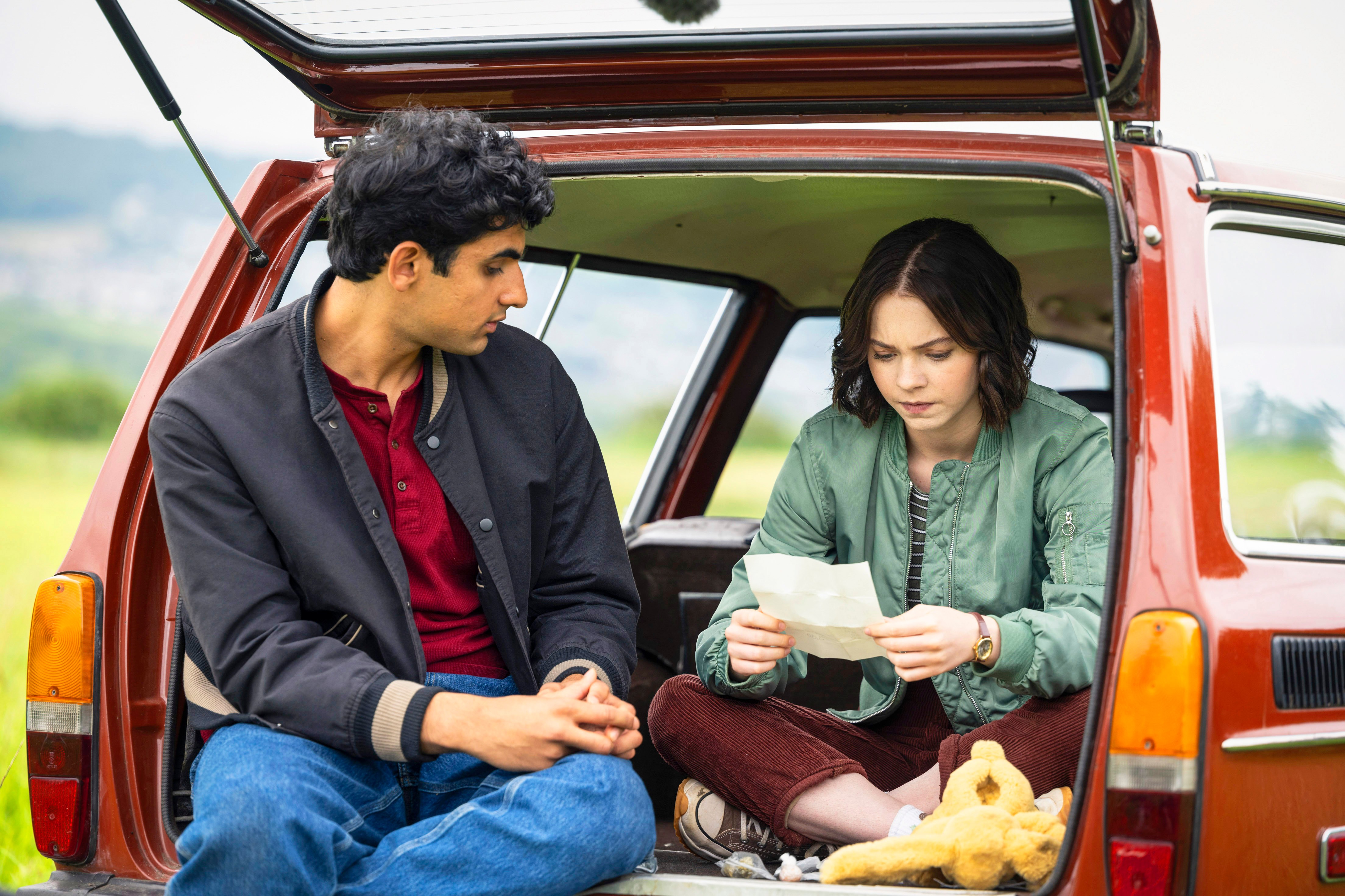 Ravi and Pip sat in the open boot of a car, looking at a piece of paper