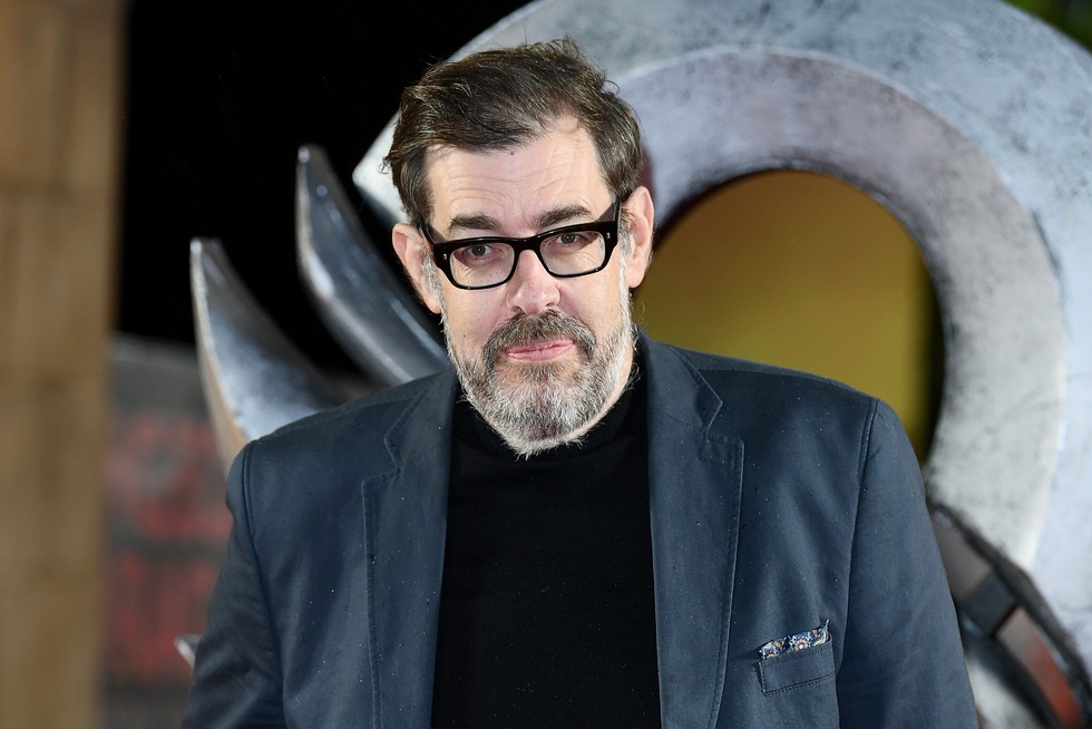 Richard Osman wearing a blue suit jacket and back jumper underneath. He is looking at the camera.