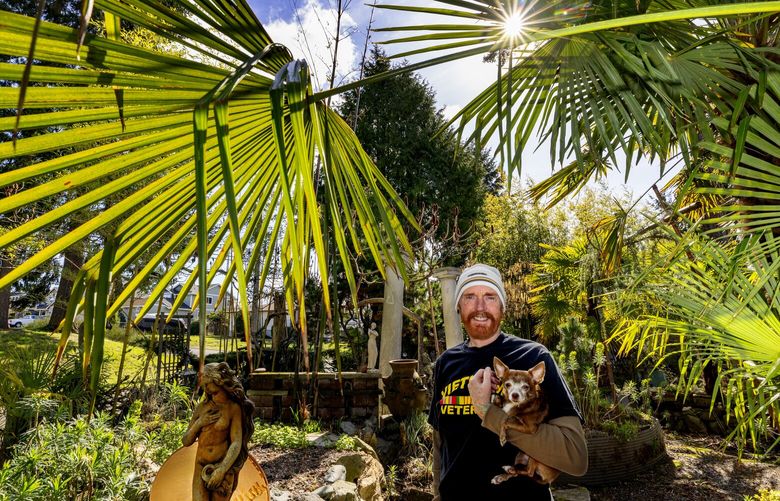 Michael Adcock holds Boopa in his tropical garden which is always a work-in-progress and wraps around the house from front yard to back, Wednesday, March 13, 2024 in Tacoma. “When I moved (here) in 1978, it was a lawn,” says Adcock.