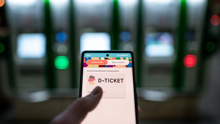 Ein Fahrgast hält ein Smartphone mit einem digitalen Deutschlandticket an einer U-Bahnstation in der Hand.