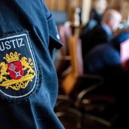 Ein Justizbeamter steht im Gerichtssaal, während der Angeklagte vor Prozessbeginn sein Gesicht hinter einer Mappe verbirgt. 