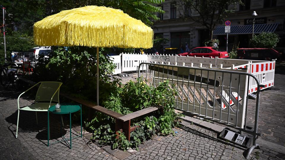 Ein Sonnenschirm und Stühle stehen vor einem mit rot-weissen Absperrungen umzäunten Parkplatz.