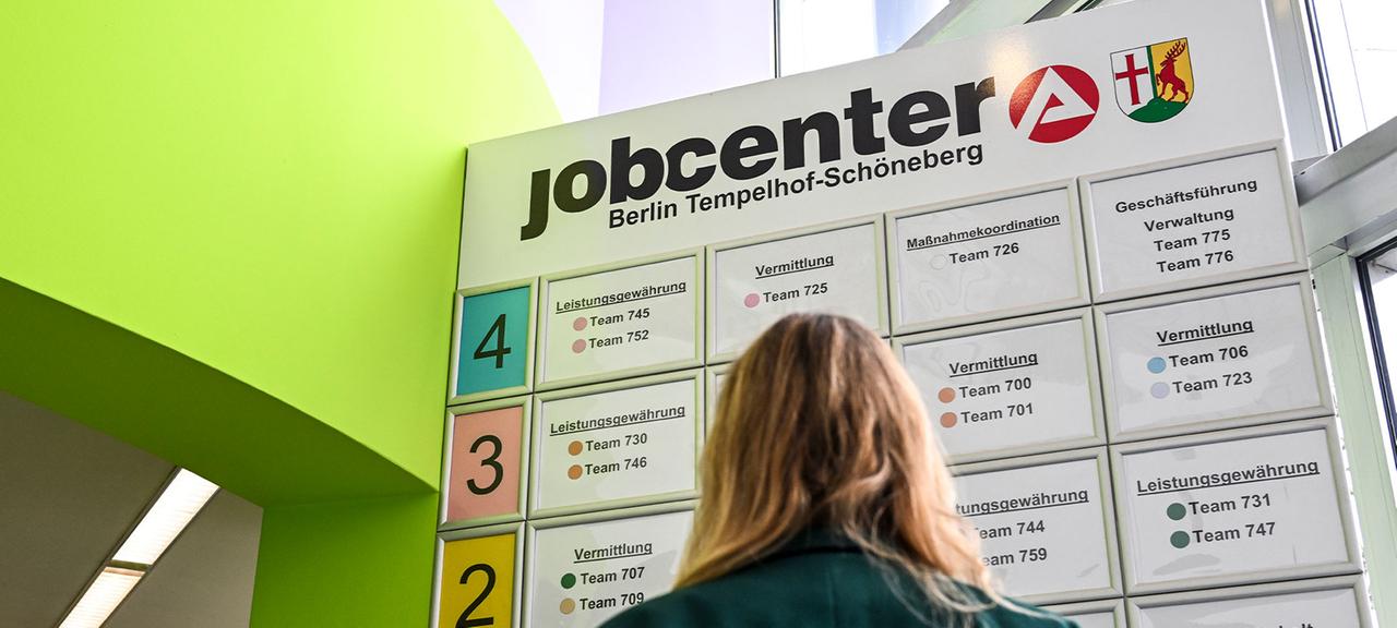 Eine Frau steht vor einer Übersicht der verschiedenen Angebote und der Leistungsgewährung im Jobcenter Tempelhof-Schöneberg. 