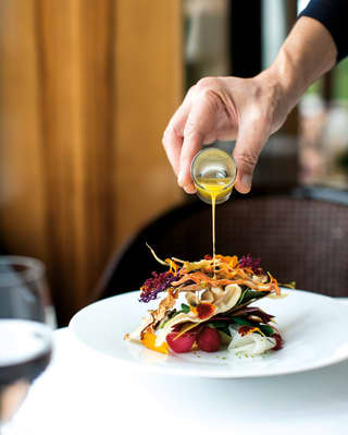 A waiter pours a small glass of dressing over an artistic tower of colourful salad arranged on a large white plate