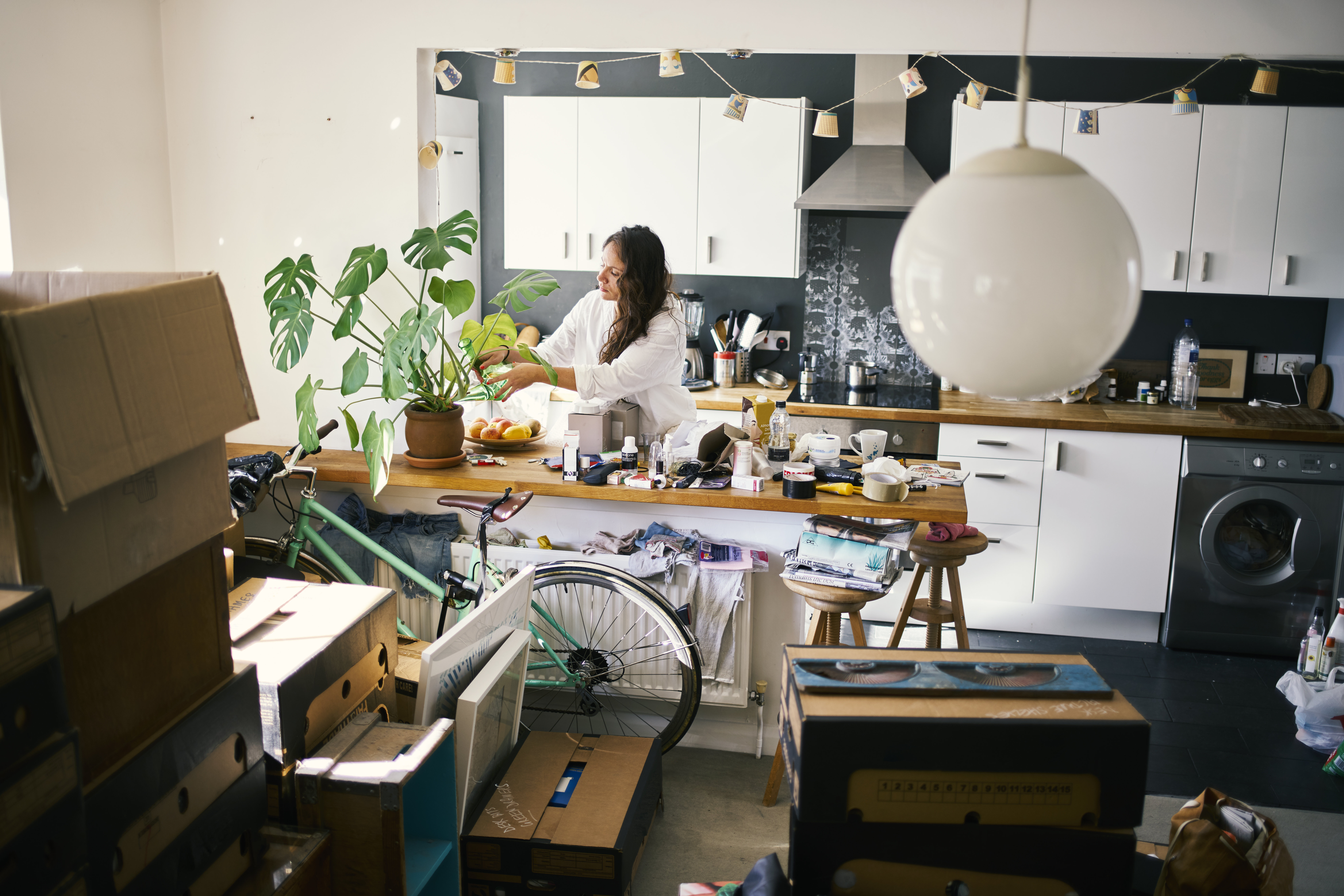 A person in a cluttered kitchen surrounded by moving boxes and a bicycle waters a houseplant on a counter filled with various items