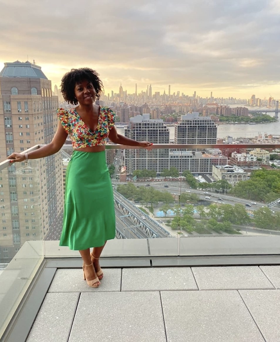 The model wears the green silk maxi while standing on an open patio in front of a skyline view