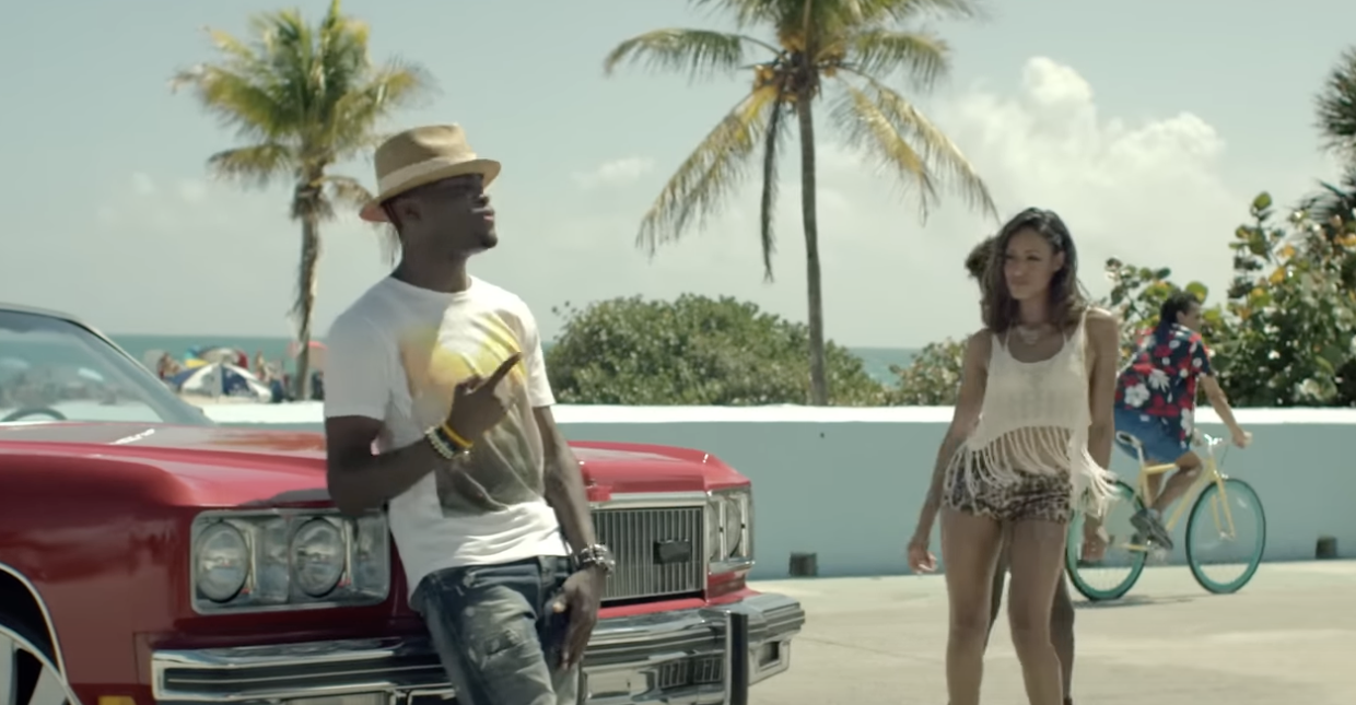 Two people stand by a classic car near the beach; man holds a football, woman in a fringed outfit