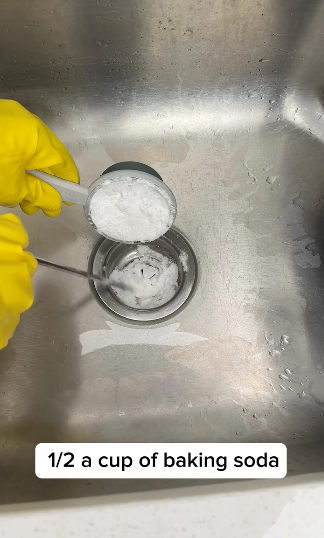 Hand in glove pouring baking soda into a cup in a sink, with caption &quot;1/2 a cup of baking soda.&quot;