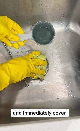 Hands wearing yellow gloves mix a substance in a cup placed in a sink with text &quot;and immediately cover&quot;