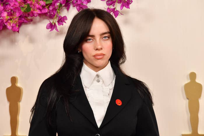 Billie Eilish posing in a black blazer and white shirt with a red accessory on the lapel at an event