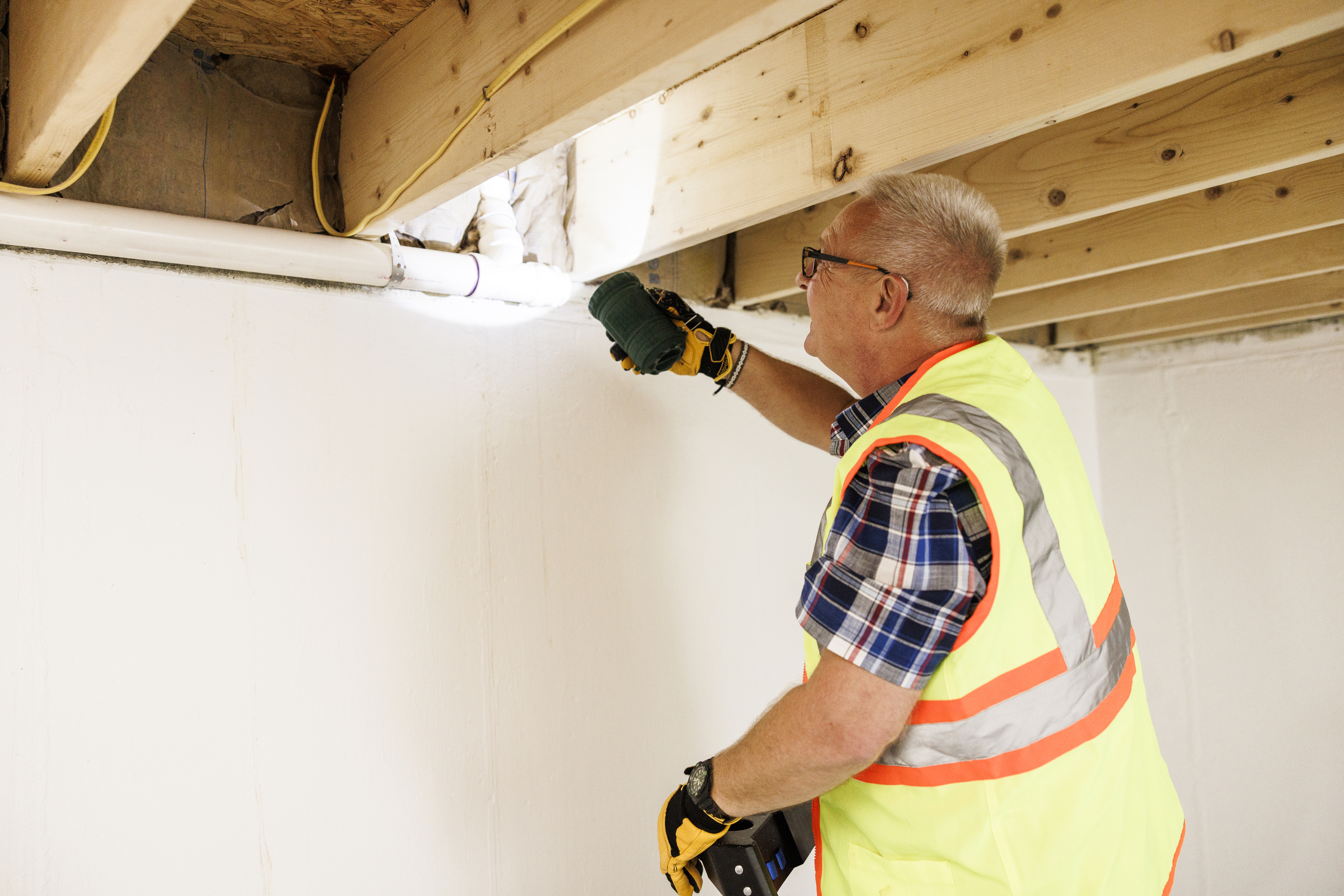 A worker inspects piping in a ceiling space using a flashlight