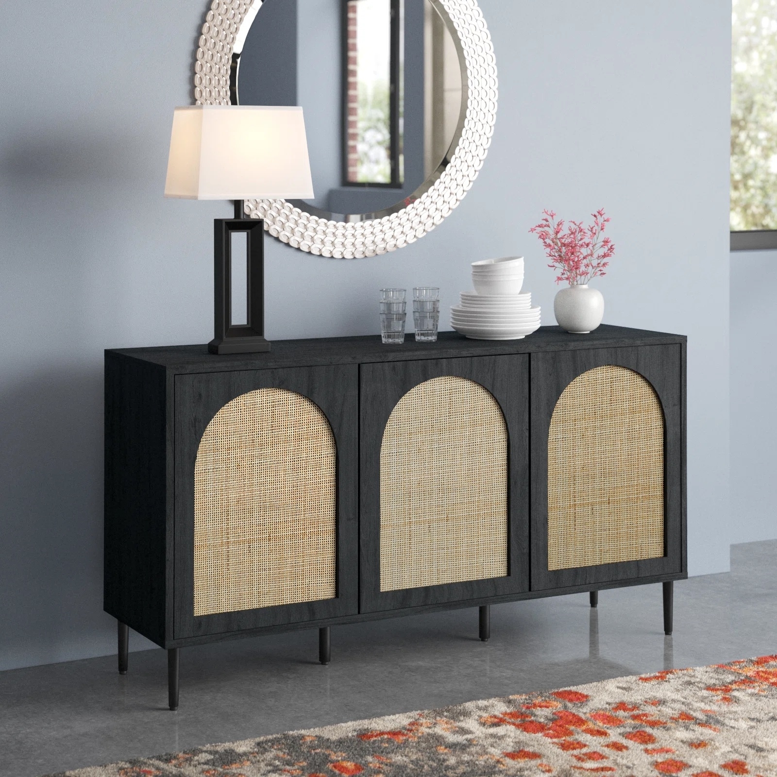 A modern sideboard with three arched wicker-front cabinets, a table lamp, a round mirror, stacked books, two drinking glasses, a flower vase, and a window in the background