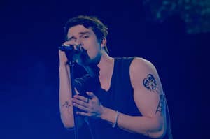 Nicholas Galitzine holding a microphone passionately performing on stage, wearing a sleeveless shirt, displaying tattoos on both arms