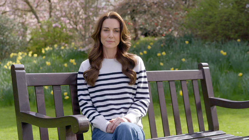 Kate Middleton sitting on a wooden bench in a garden with daffodils in the background, wearing a striped sweater and jeans