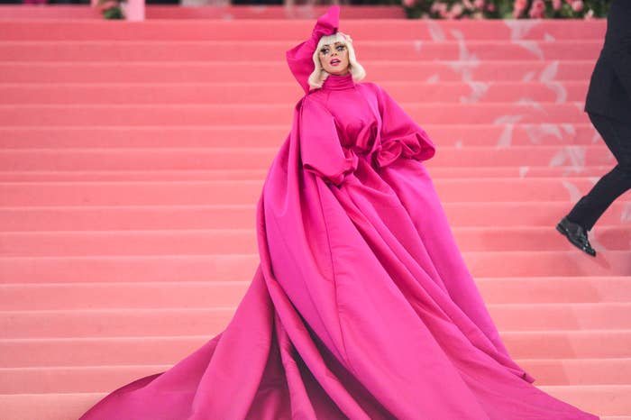 Lady Gaga poses on stairs in a voluminous, flowing gown with a high neckline and oversized sleeves, accessorized with a matching bow headpiece
