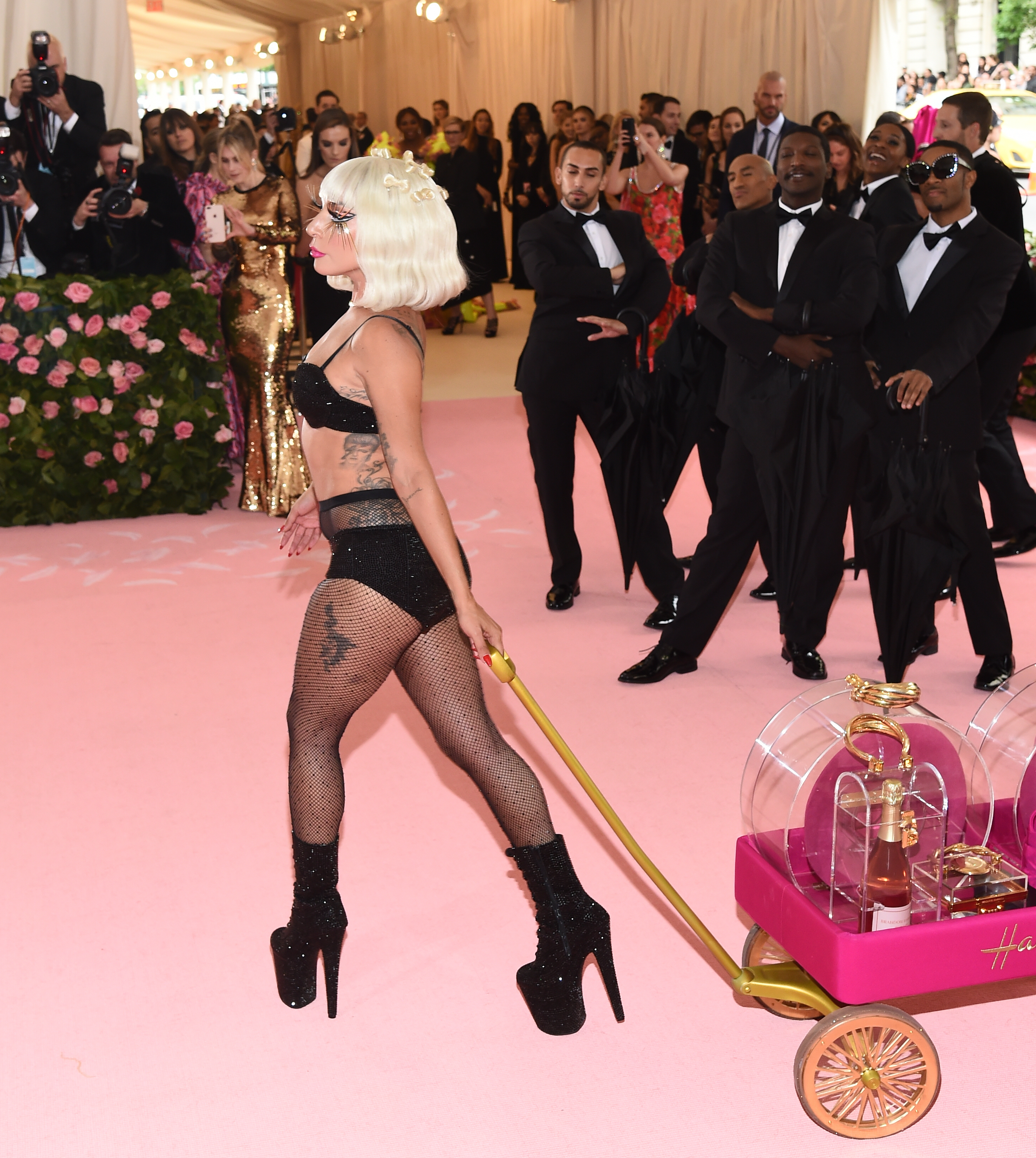 Lady Gaga walking on the carpet wearing a bra and panties with platform heels, pulling a cart with large perfume bottles