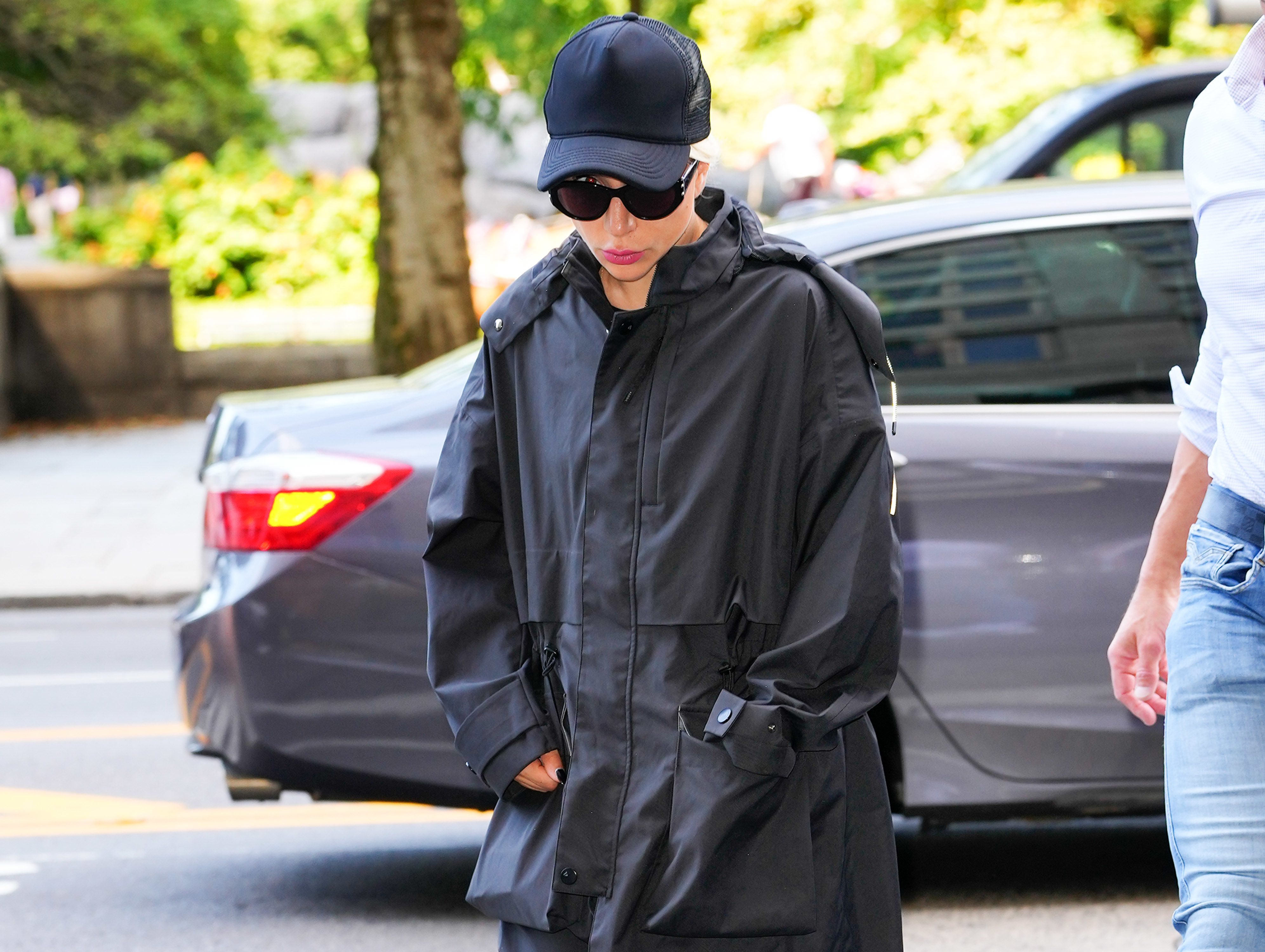 Lady Gaga wearing a cap, sunglasses, and a hooded jacket walks on a city street near a car, with another person partially visible beside them