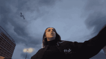 I don&#x27;t know who this person is. The image shows a person in motion looking up at the sky, wearing a dark jacket that says &quot;Proud.&quot; Buildings and streetlights are in the background