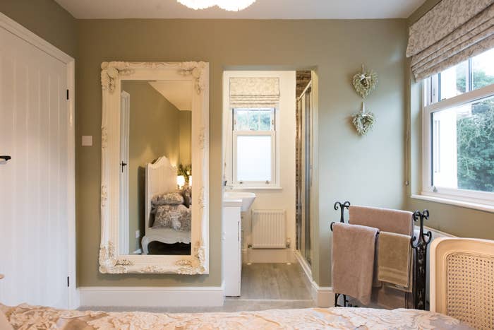 A cozy bedroom with a large ornate mirror, a bed with a floral comforter, a window, a towel rack, and a doorway leading to a sink area