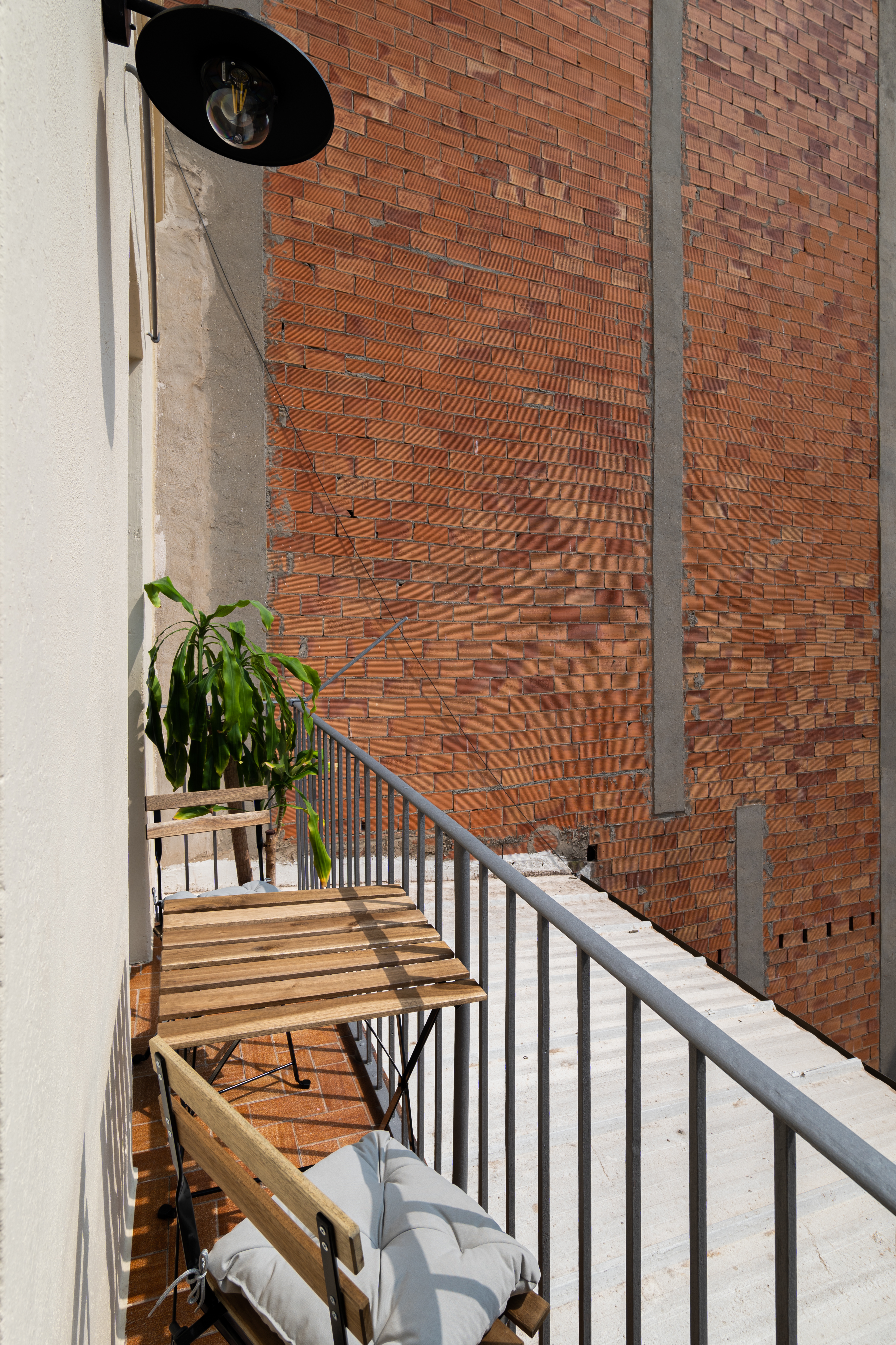 A small balcony with wooden furniture and a potted plant, flanked by a brick wall and railing