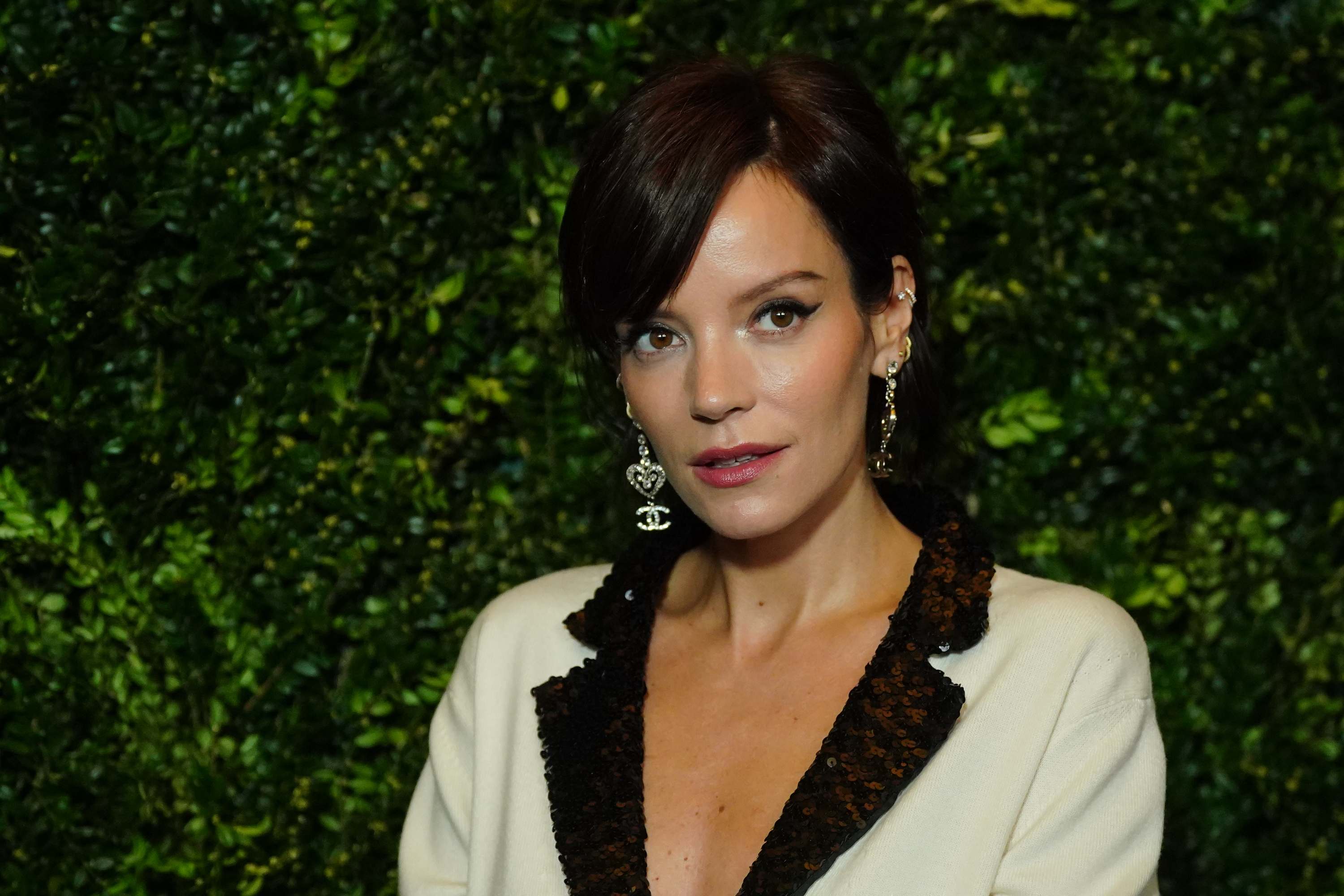 Lily Allen poses in a blazer with dark lapels in front of a leafy backdrop. She has short hair and wears dangling earrings