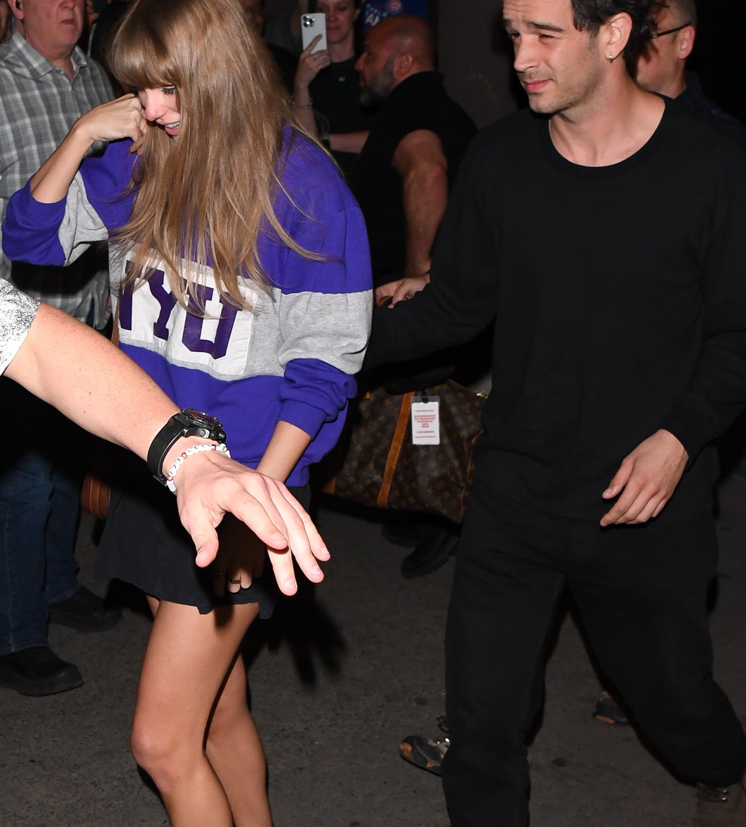 Taylor Swift is wearing an NYU sweatshirt, a skirt, an sneakers. She is holding hands with Matty Healy, who is wearing a dark outfit. They are walking outside, surrounded by people