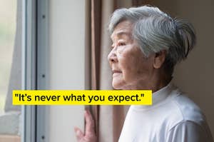 Elderly woman with short hair, in a white turtleneck, looking contemplatively out a window, hand resting on the curtain