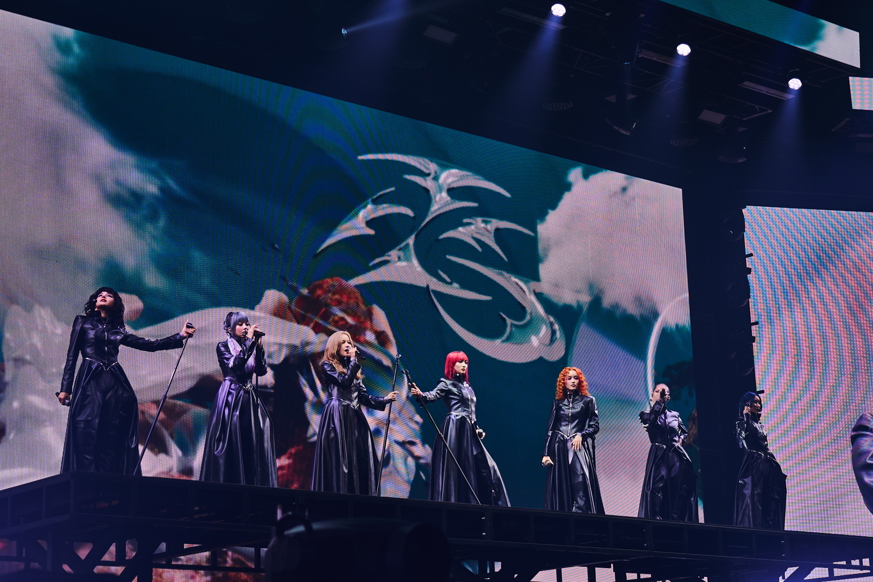 Performers on stage in long, black outfits during a theatrical production with a large, dramatic backdrop