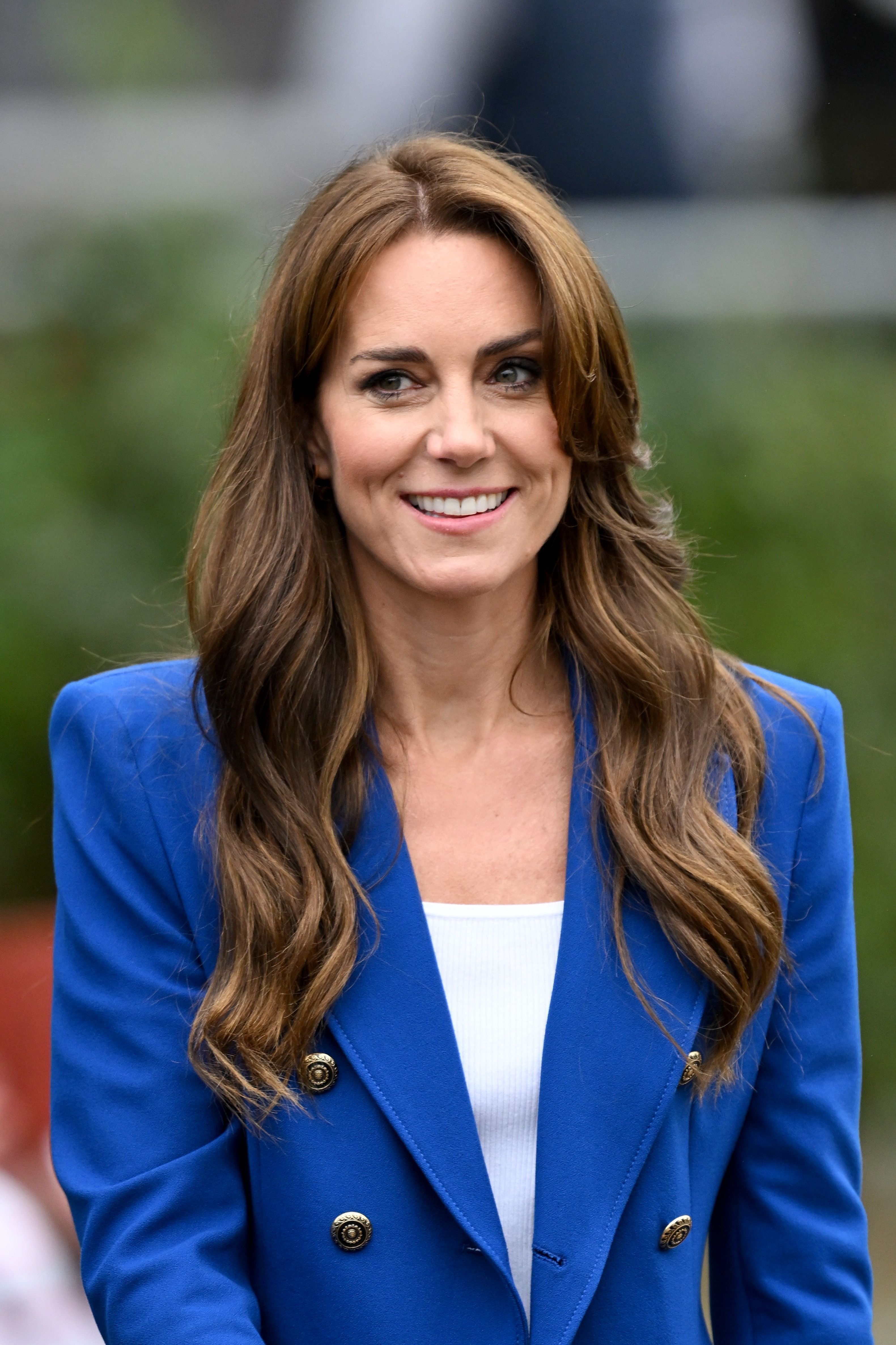 Kate Middleton smiles in an outdoor setting, wearing a tailored blazer with gold buttons over a white top