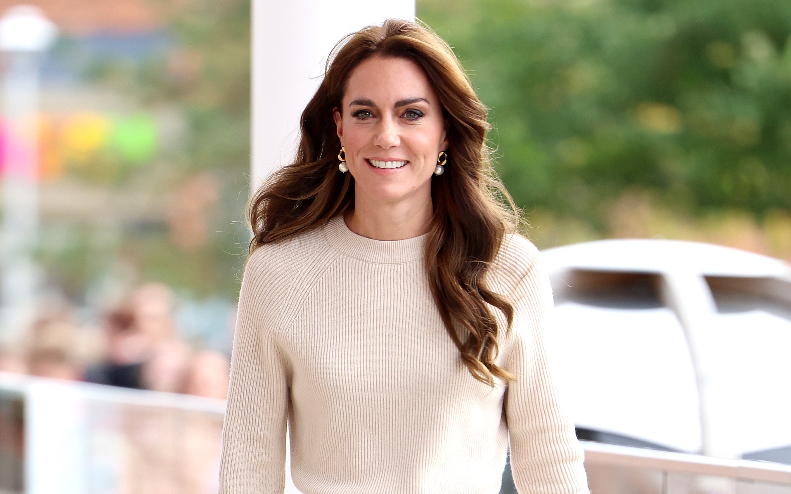 Kate Middleton, smiling, wearing a beige sweater with pearl earrings, walking outdoors