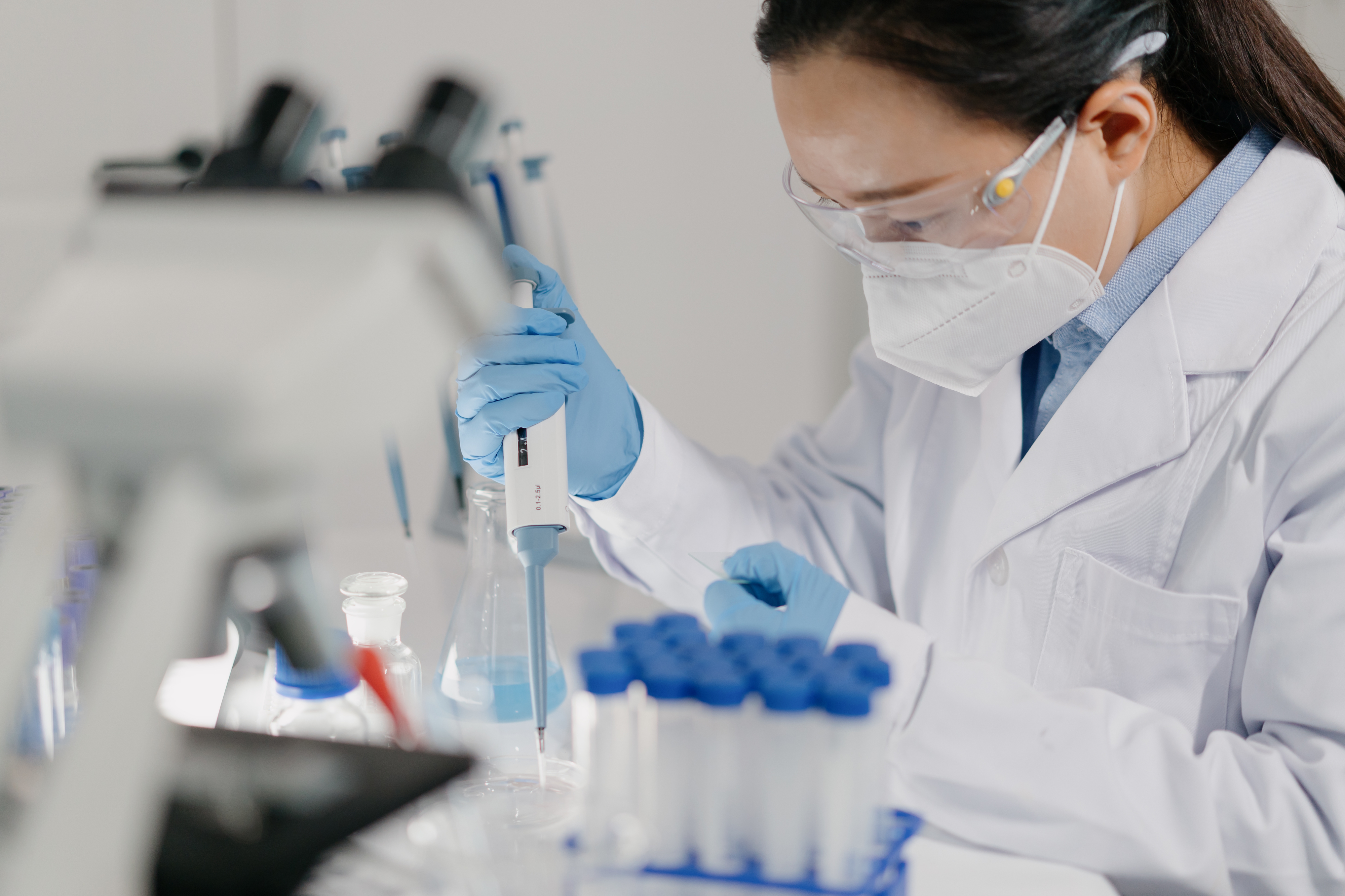 A scientist using a pipette in a lab setting, wearing a lab coat, gloves, and a mask, focused on an experiment