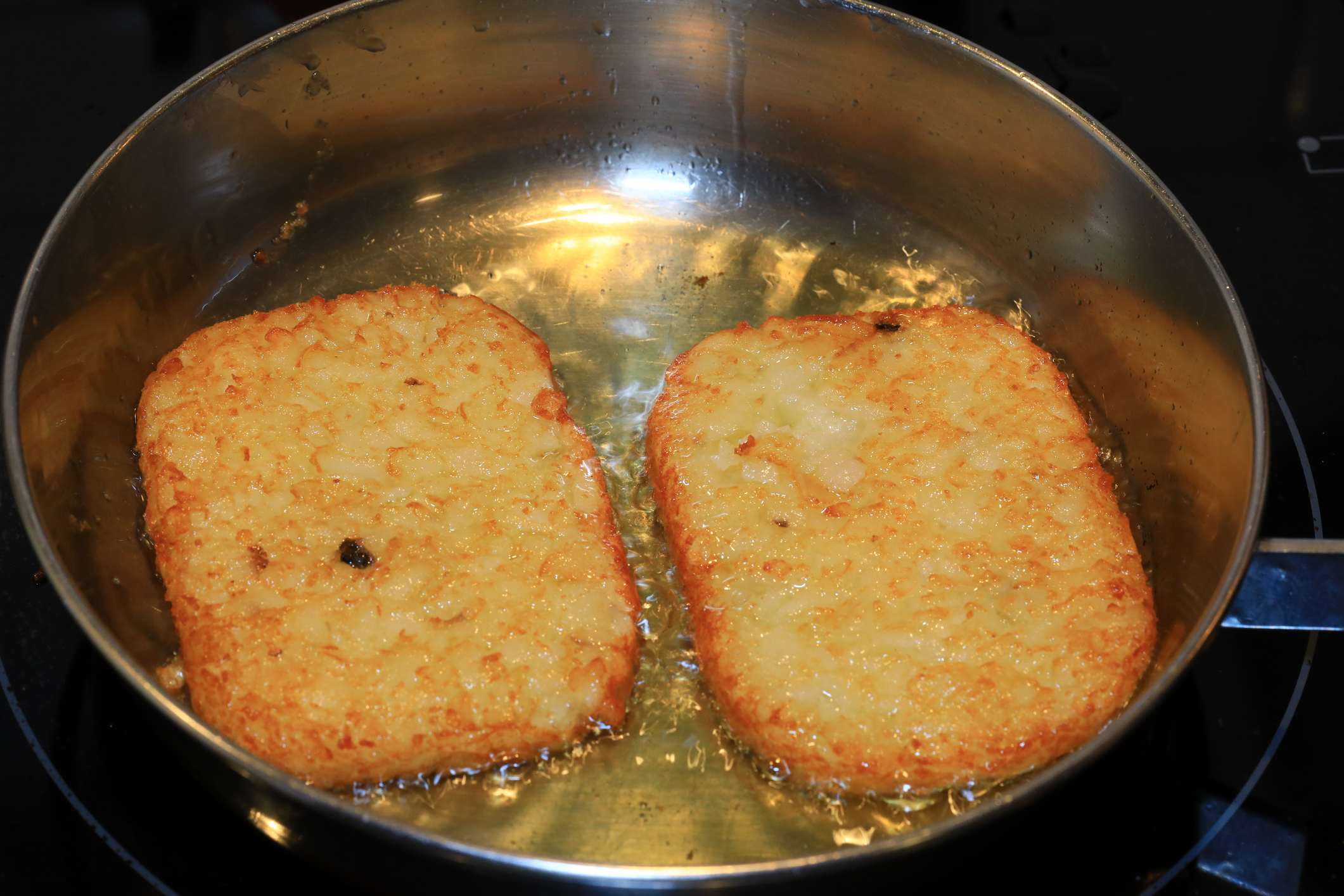 Two hash browns frying in a pan with hot oil