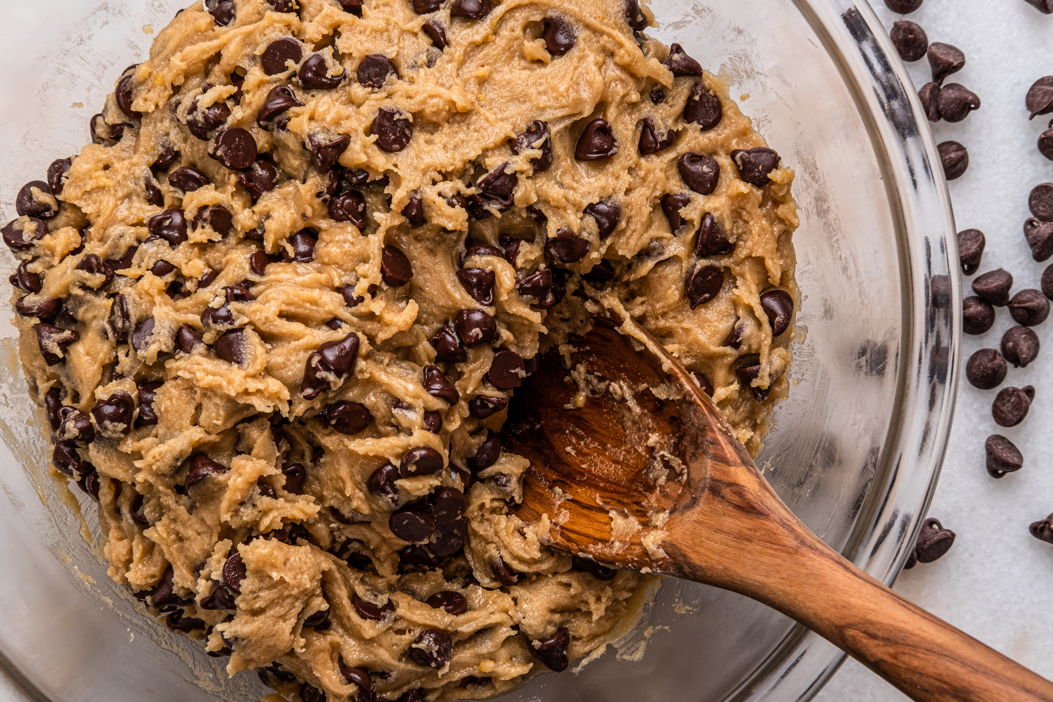 A glass bowl filled with chocolate chip cookie dough, being stirred with a wooden spoon. Chocolate chips are scattered around the bowl