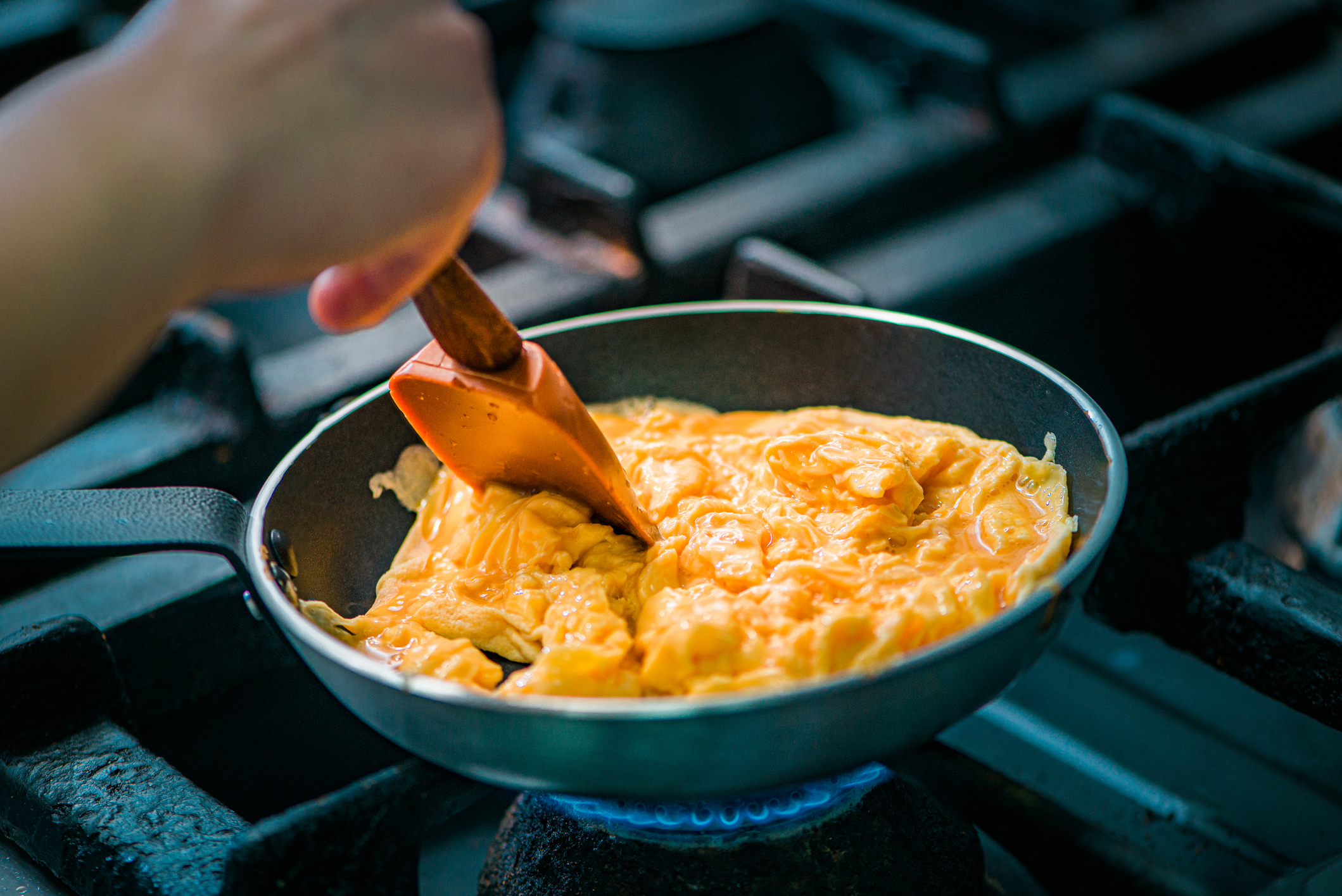 A person is cooking scrambled eggs in a pan on a stove, using a wooden spatula to stir them