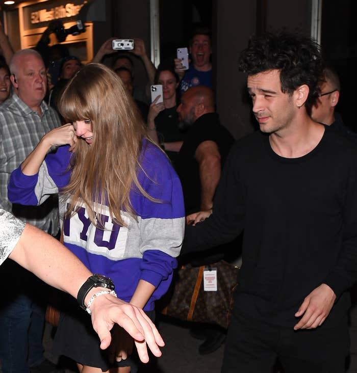 Taylor Swift and Matty Healy smiling while walking together, surrounded by fans and photographers. Taylor is wearing a &#x27;NYU&#x27; sweatshirt and skirt