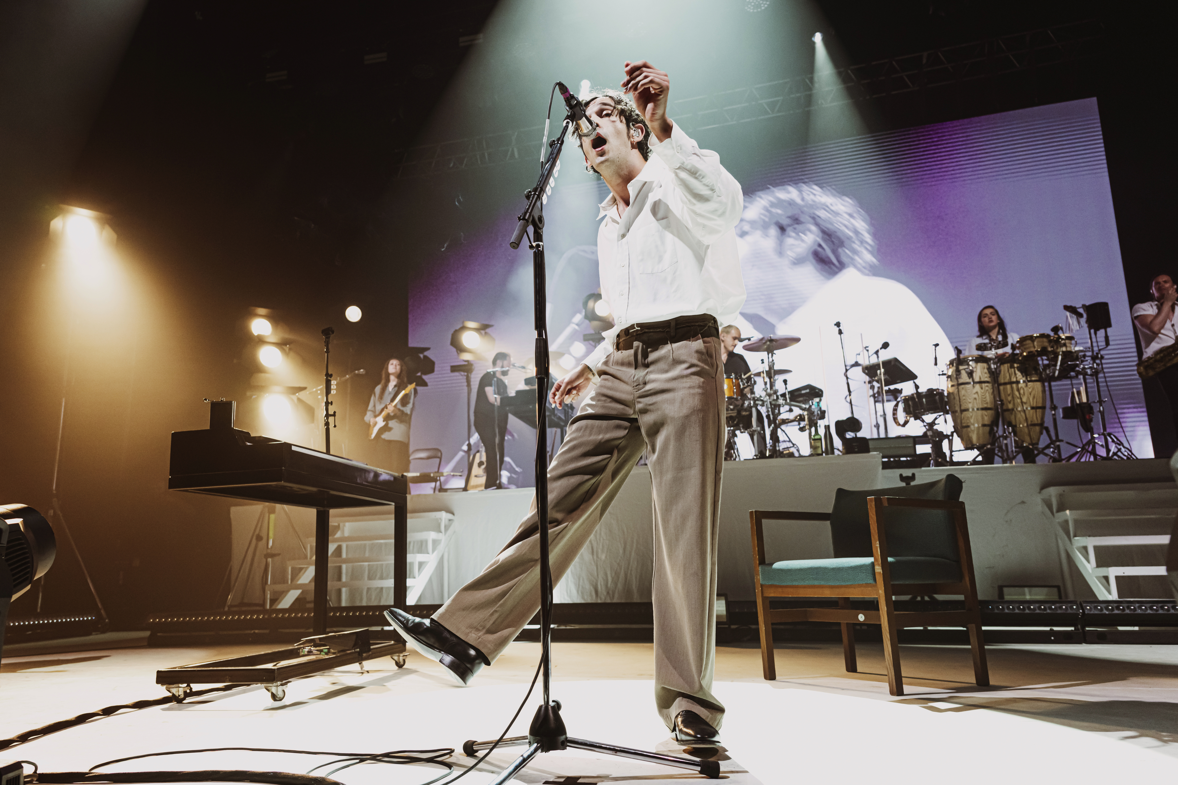 Matty Healy performs energetically on stage, wearing a dress shirt and slacks, with a band and musical instruments in the background