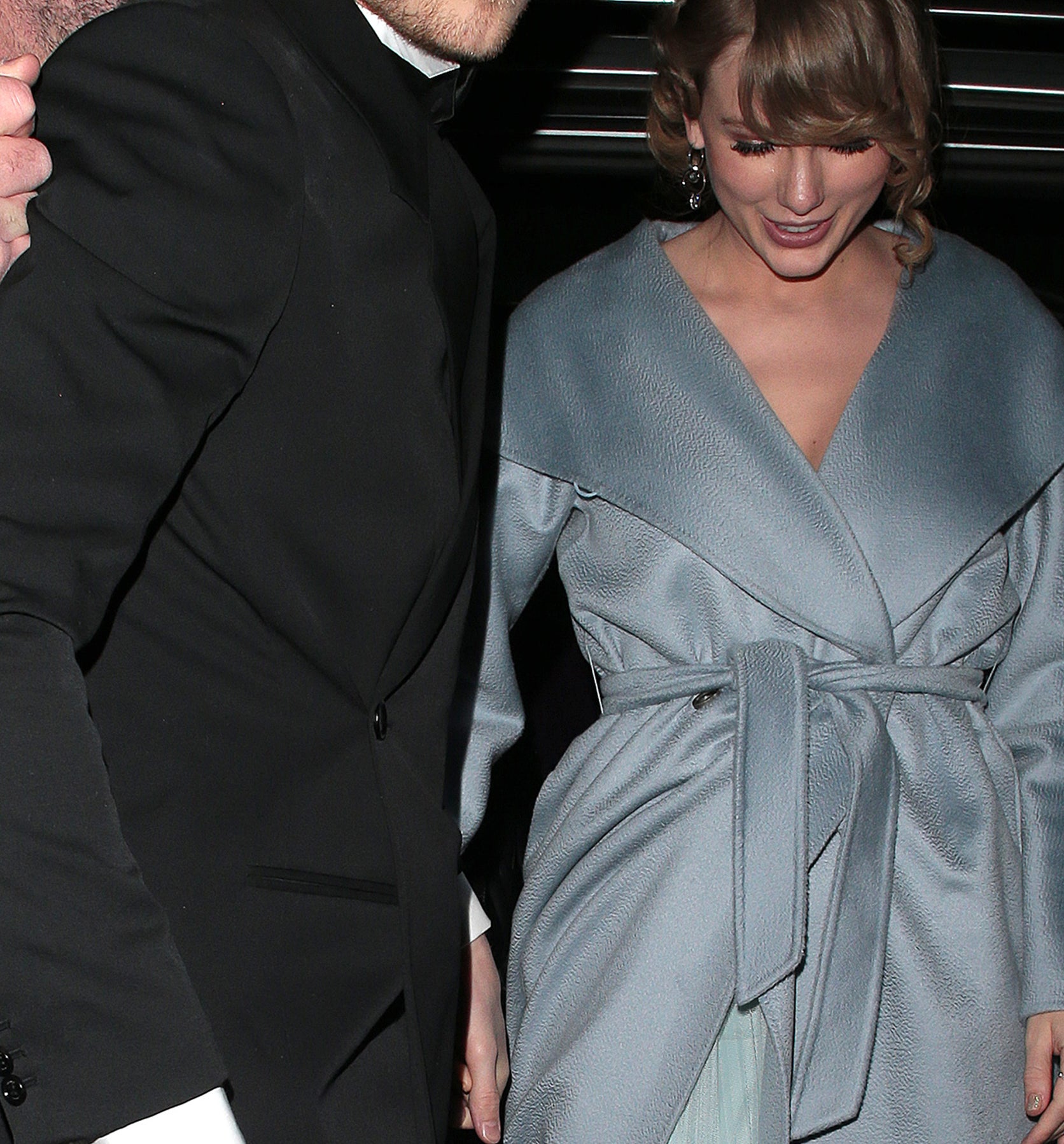 Joe Alwyn in a suit and Taylor Swift in a long coat are seen walking together, guided by an assistant holding an umbrella