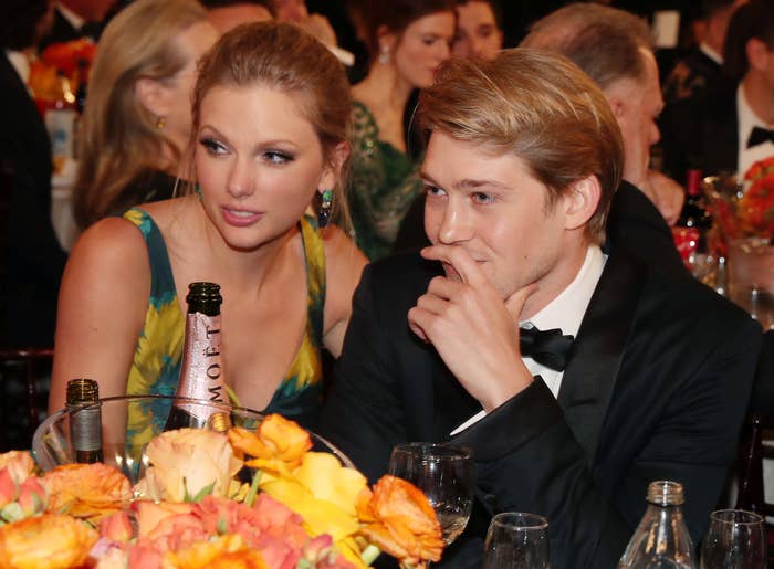 Taylor Swift in a dress and Joe Alwyn in a tuxedo sit at a formal event table with flowers
