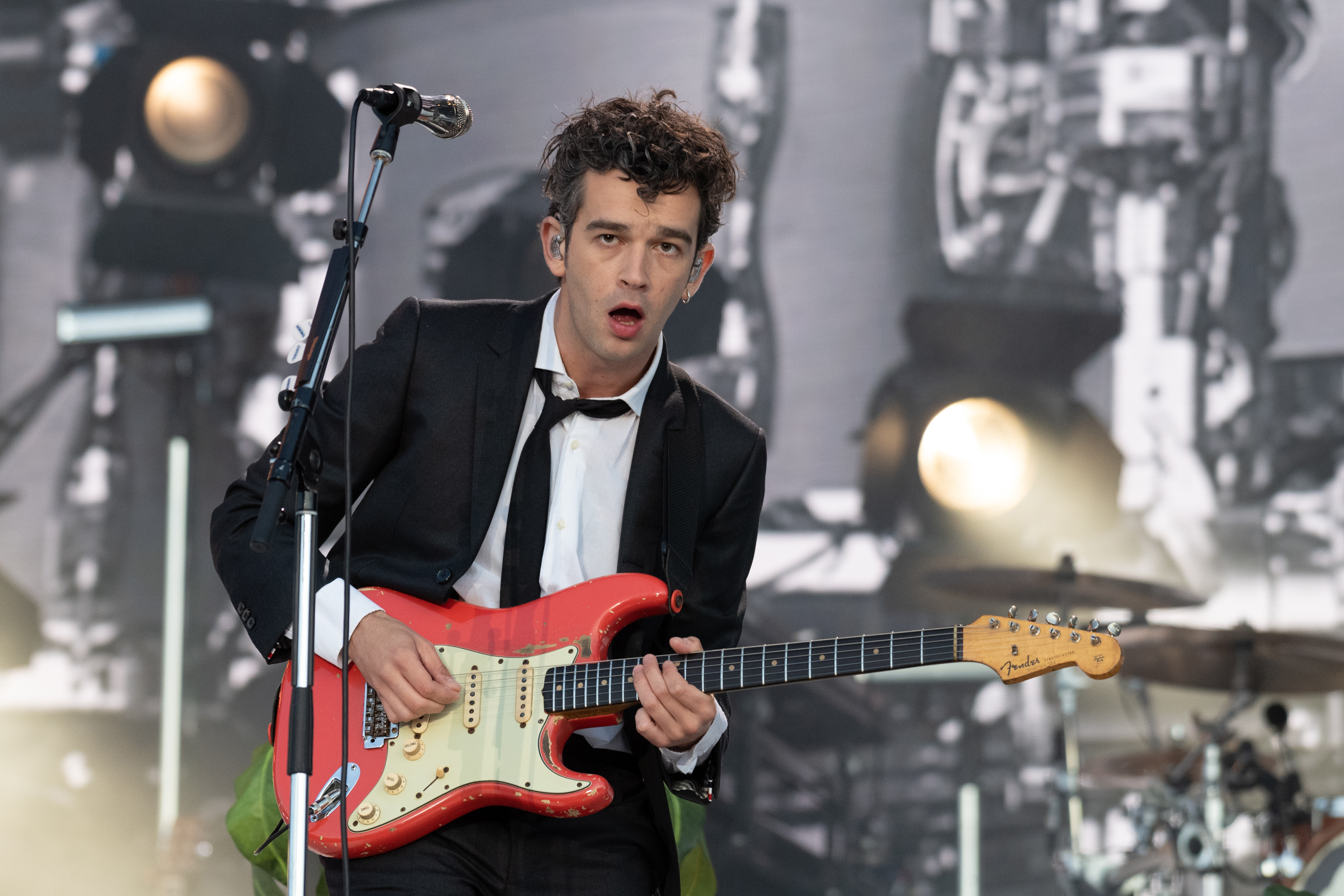 Matty Healy, wearing a black suit and white shirt, performs on stage with a red electric guitar