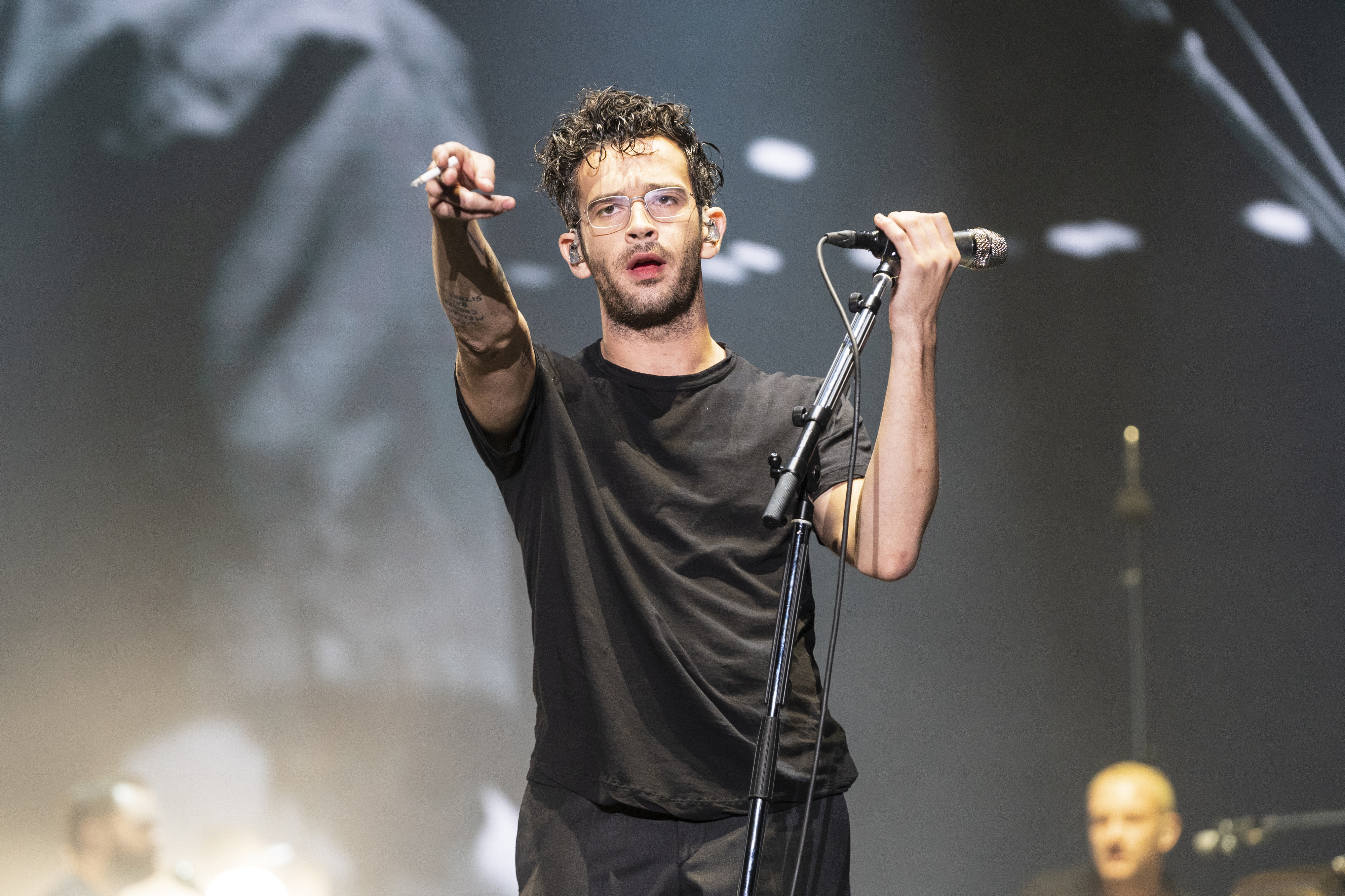 Matty Healy performs on stage, wearing a casual black outfit and holding a microphone, with a band member visible in the background