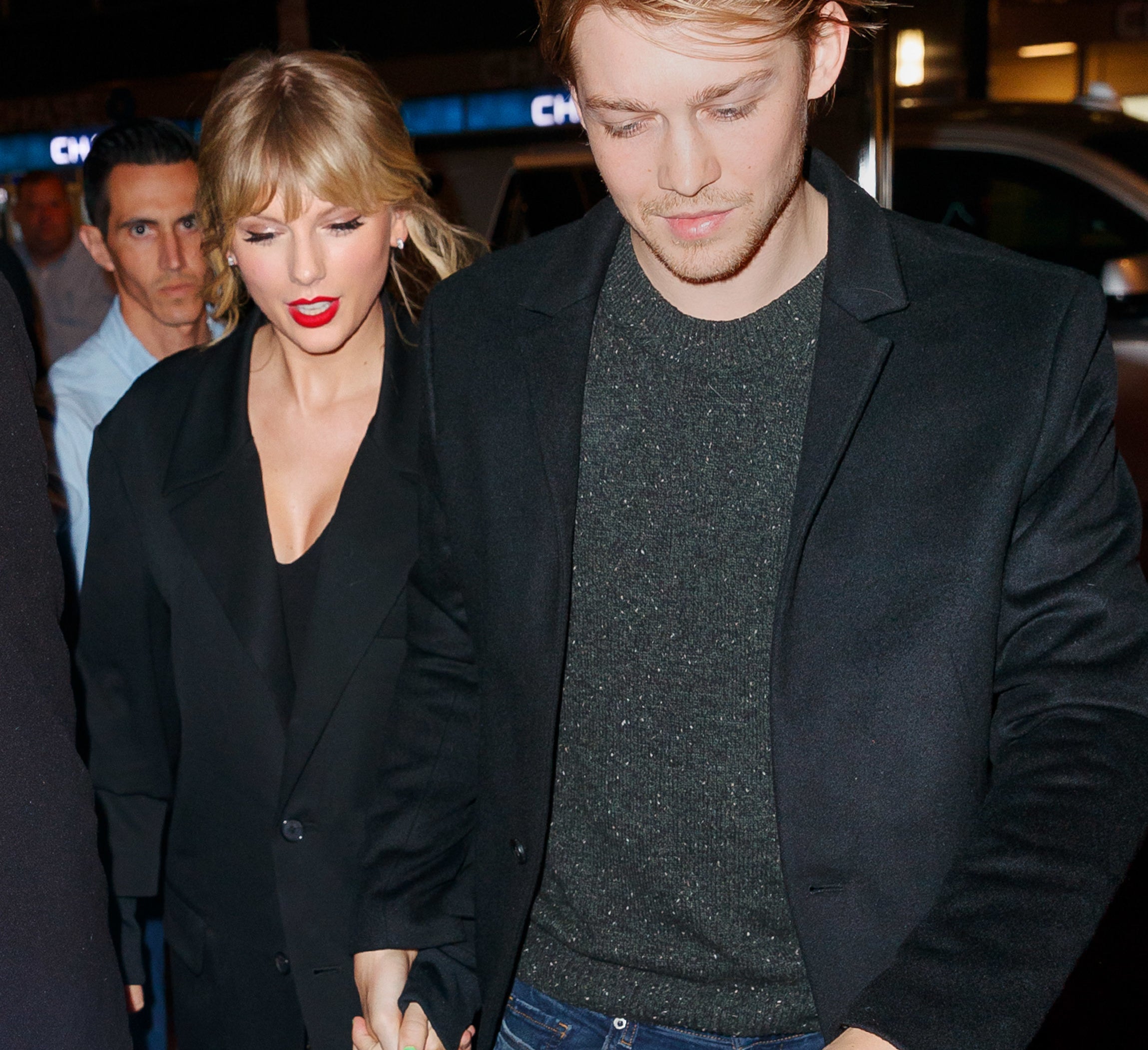 Taylor Swift and Joe Alwyn are holding hands, walking together. Taylor is wearing a blazer and patterned pants. Joe is wearing a dark coat and jeans