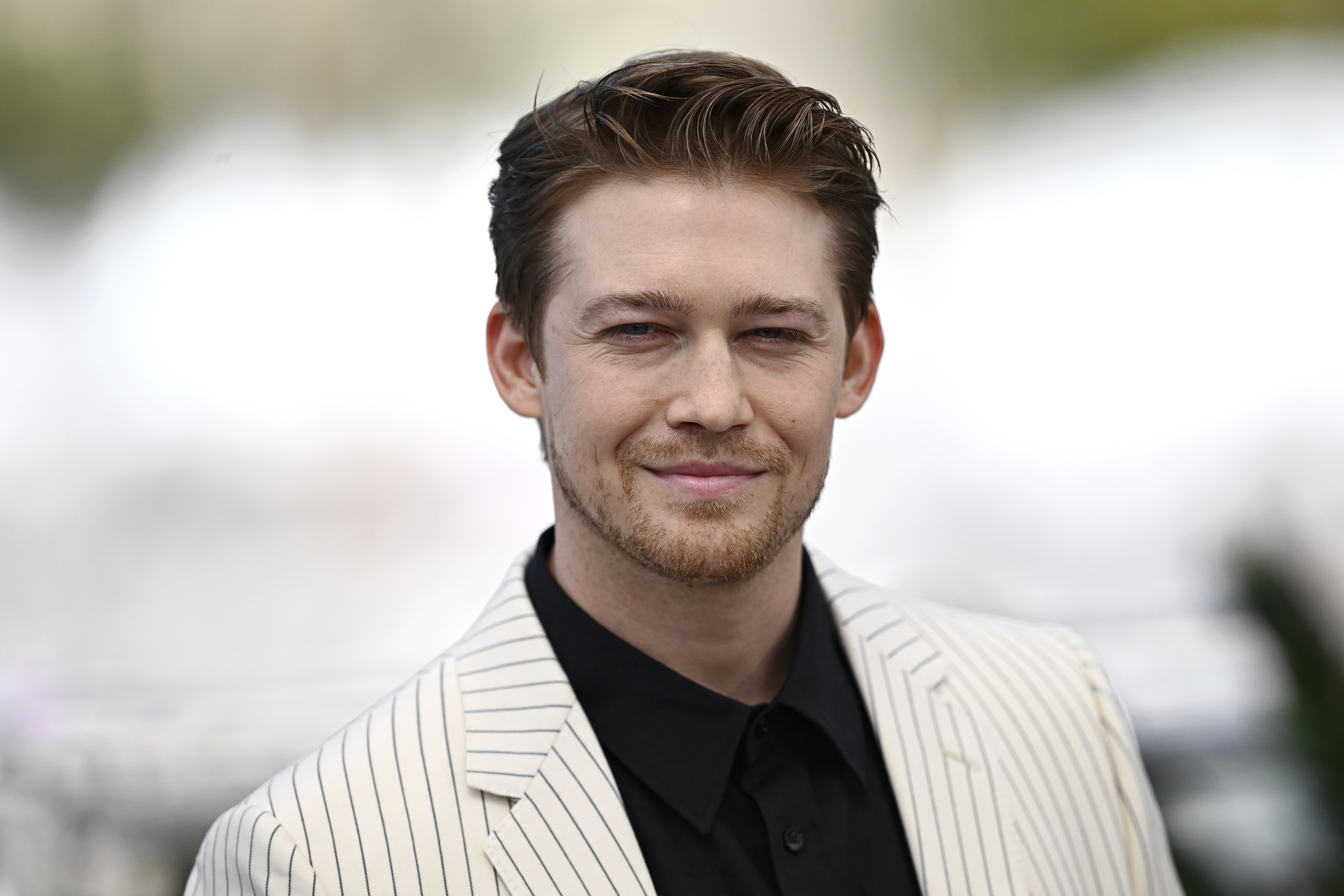 Joe Alwyn is wearing a light striped suit jacket and a dark shirt, posing and smiling