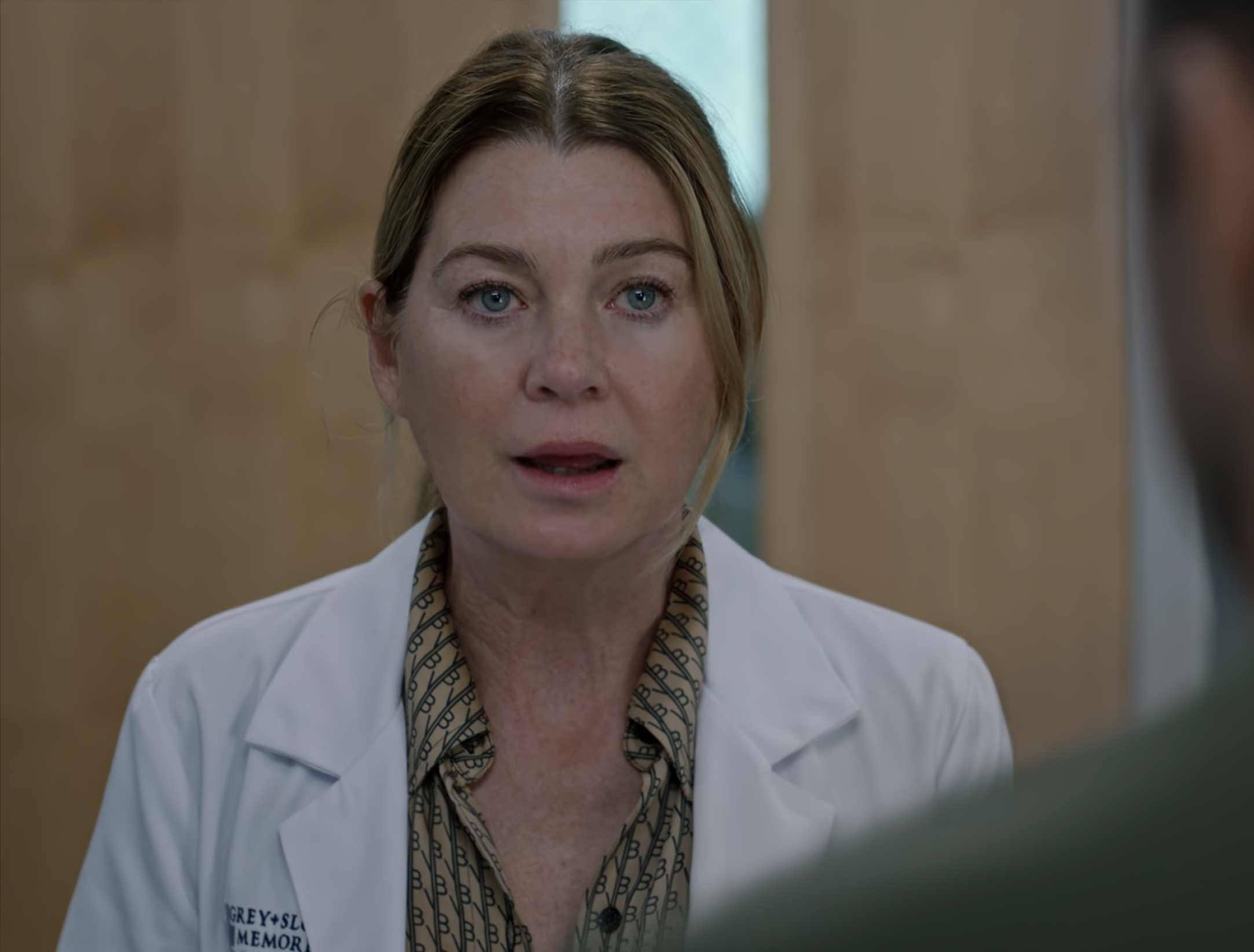 Ellen Pompeo, as Dr. Meredith Grey, speaks to someone off-camera in a scene. She is wearing a doctor&#x27;s coat over patterned clothing