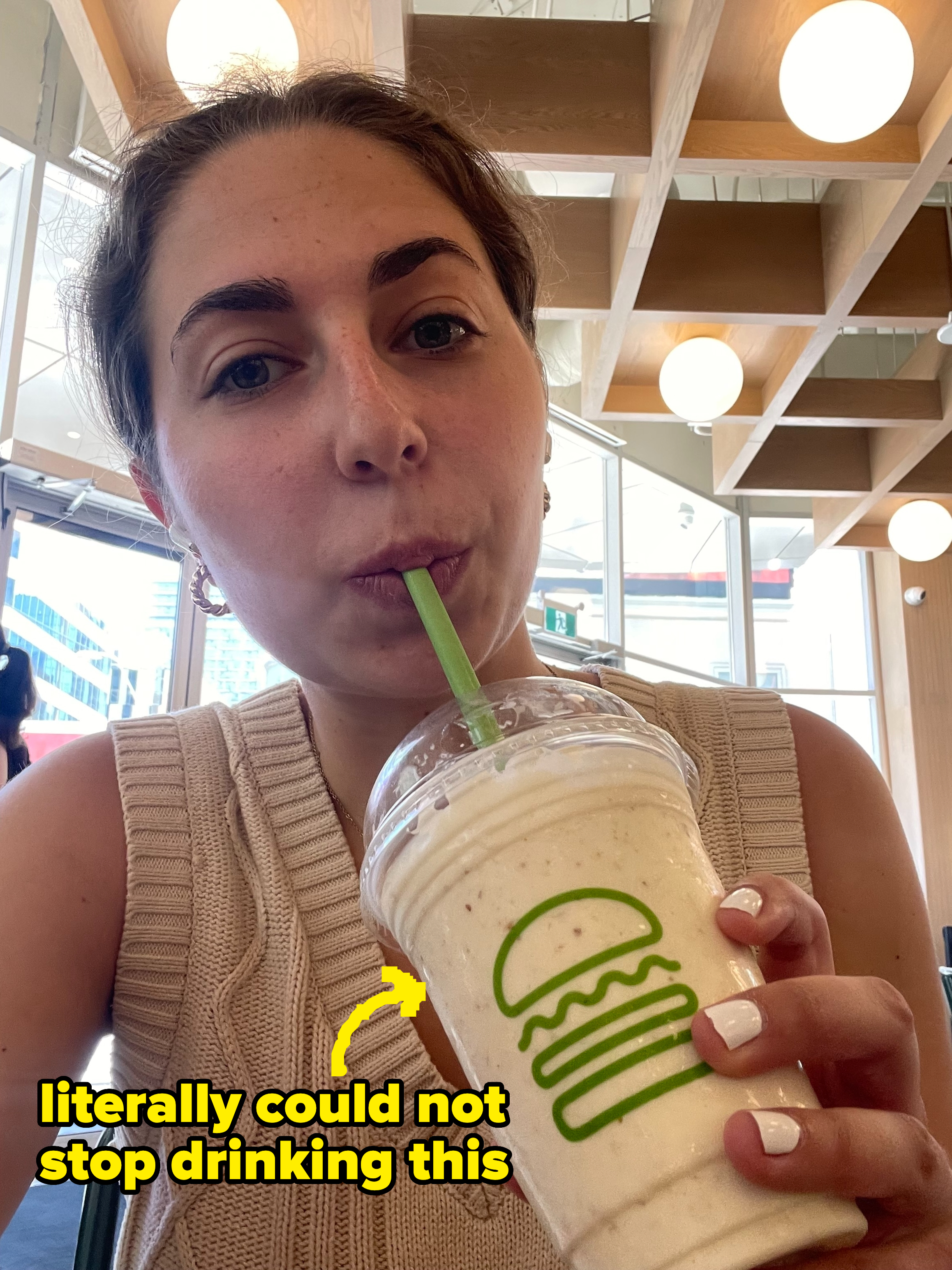 Person drinking a milkshake with a green straw inside a fast-food restaurant. Cup features a logo of a burger with a person wearing a sleeveless knit top